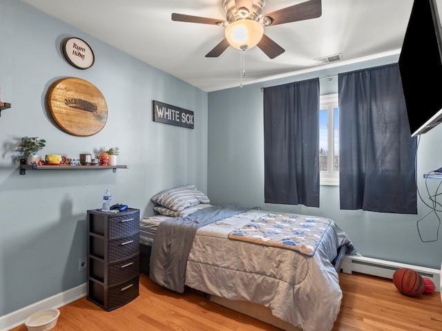 bedroom featuring ceiling fan, a baseboard heating unit, wood finished floors, visible vents, and baseboards