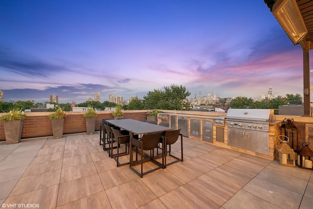 patio terrace at dusk featuring a view of city, outdoor dining area, an outdoor kitchen, and a grill