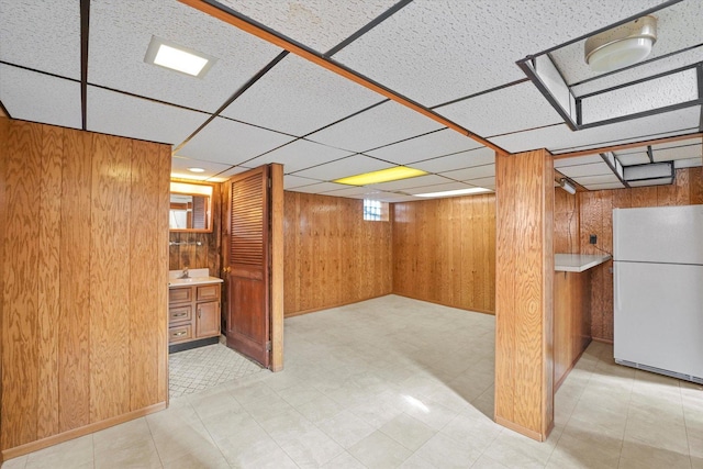 below grade area featuring tile patterned floors, freestanding refrigerator, a paneled ceiling, wood walls, and a sink