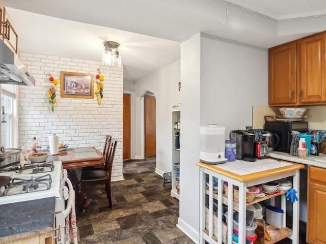 kitchen featuring arched walkways, brown cabinetry, brick wall, stone finish floor, and ventilation hood