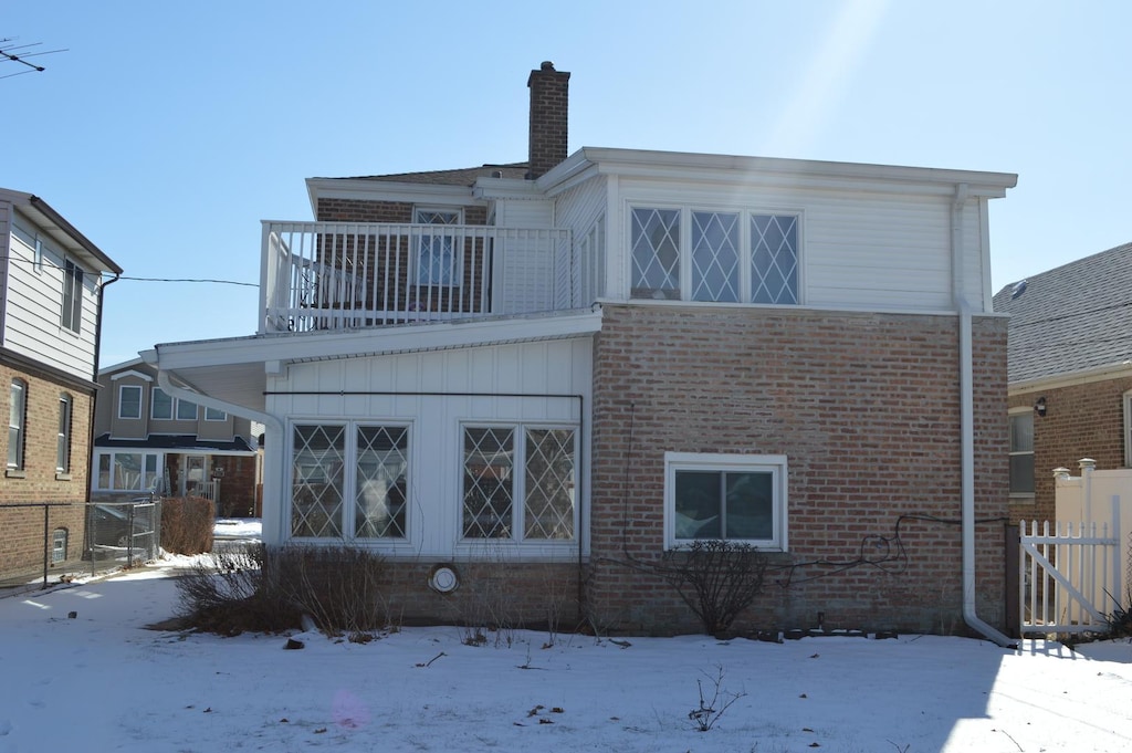 snow covered house featuring a chimney, brick siding, fence, and a balcony