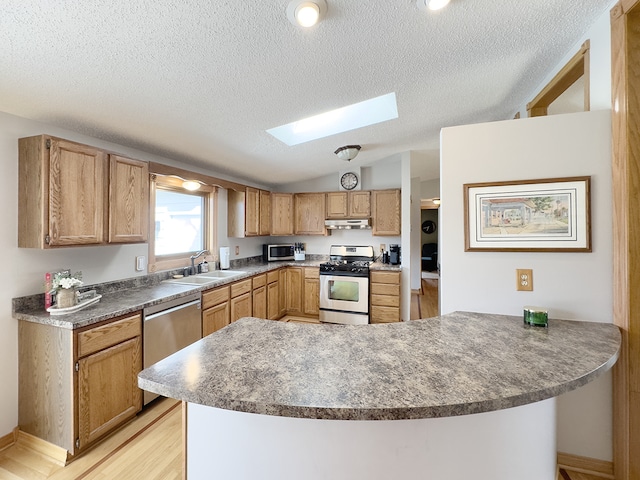 kitchen featuring under cabinet range hood, a peninsula, a sink, appliances with stainless steel finishes, and lofted ceiling with skylight