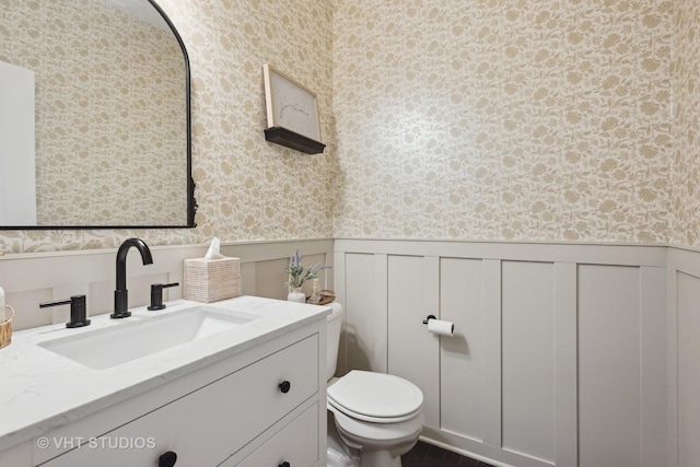 half bathroom with wainscoting, vanity, toilet, and wallpapered walls