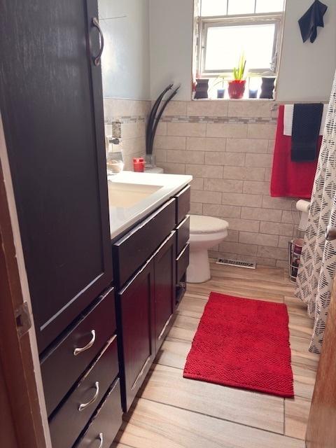 bathroom with toilet, visible vents, vanity, tile walls, and wainscoting