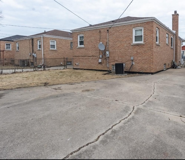 back of house featuring driveway, central AC unit, a chimney, fence, and brick siding