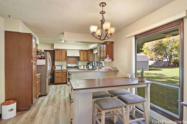 kitchen featuring brown cabinets, light wood finished floors, appliances with stainless steel finishes, a peninsula, and wall chimney exhaust hood