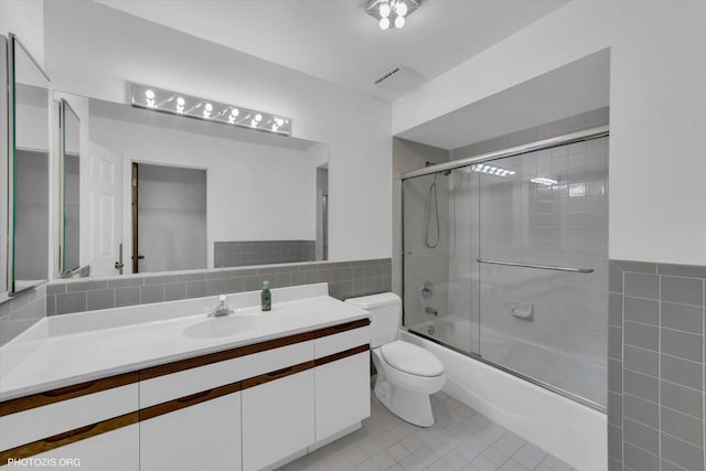 bathroom featuring tile patterned flooring, toilet, bath / shower combo with glass door, vanity, and tile walls