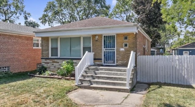 bungalow featuring brick siding, a shingled roof, covered porch, fence, and a front lawn