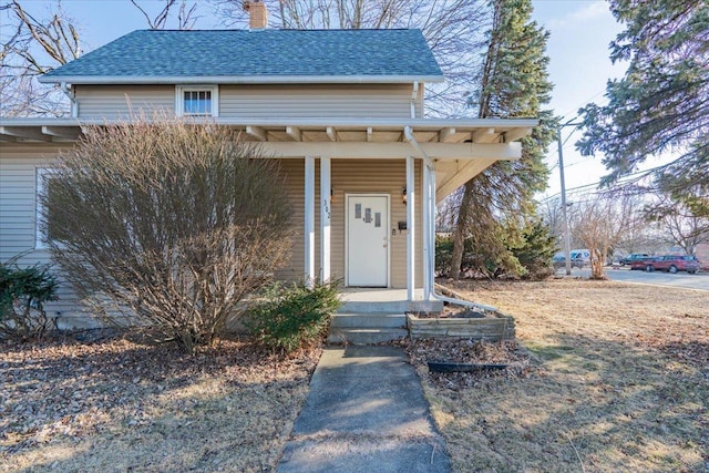 view of exterior entry featuring covered porch, a shingled roof, and a chimney