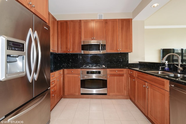 kitchen featuring stainless steel appliances, a sink, backsplash, dark stone counters, and brown cabinetry