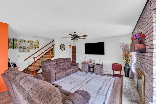 living area featuring ceiling fan, a baseboard radiator, wood finished floors, stairway, and a brick fireplace