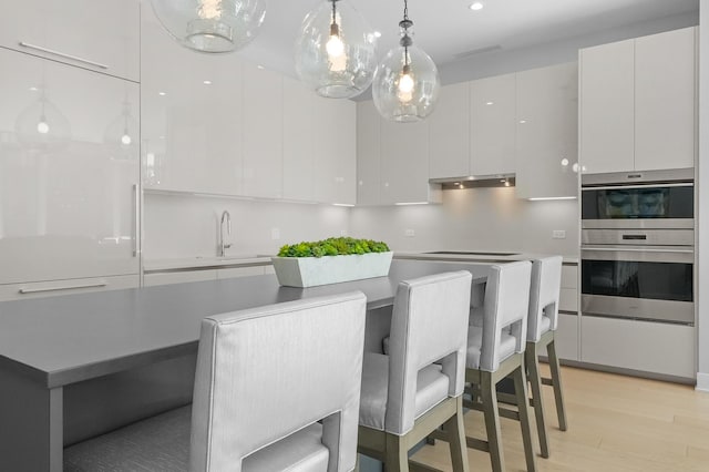 kitchen featuring double oven, under cabinet range hood, light wood finished floors, modern cabinets, and decorative light fixtures