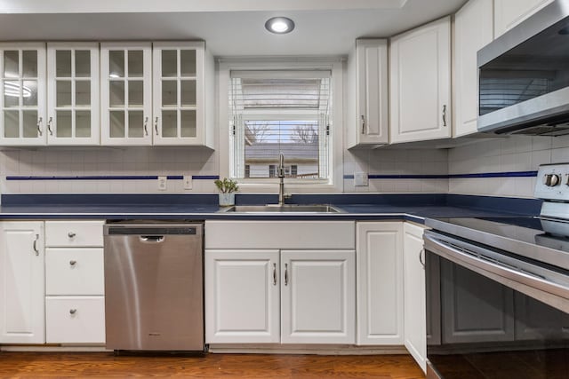 kitchen with dark countertops, glass insert cabinets, stainless steel appliances, white cabinetry, and a sink