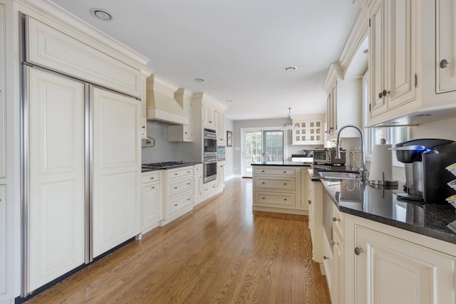 kitchen featuring stainless steel double oven, light wood-style flooring, a sink, glass insert cabinets, and custom range hood