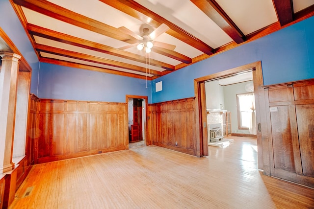 empty room with ceiling fan, a wainscoted wall, light wood-style floors, beamed ceiling, and ornate columns