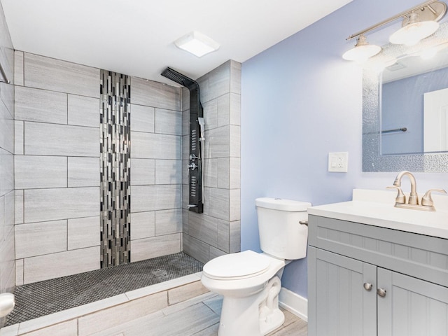 bathroom with toilet, wood finished floors, vanity, baseboards, and tiled shower