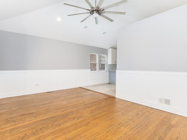 spare room with light wood-style flooring, a wainscoted wall, visible vents, a ceiling fan, and vaulted ceiling