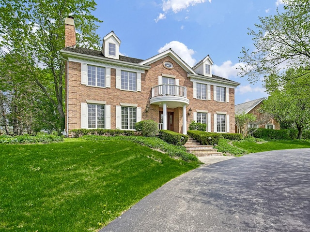 colonial-style house featuring brick siding, a front lawn, a chimney, and a balcony