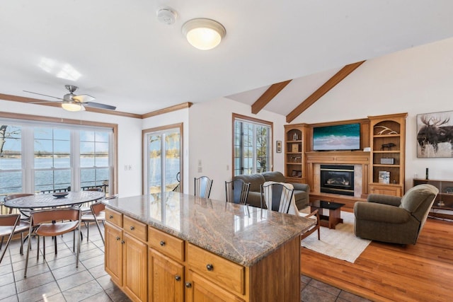 kitchen with dark stone countertops, a kitchen island, lofted ceiling, ceiling fan, and a glass covered fireplace