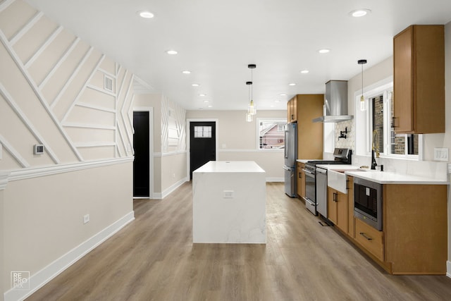 kitchen with brown cabinetry, wall chimney exhaust hood, appliances with stainless steel finishes, a center island, and light wood-type flooring