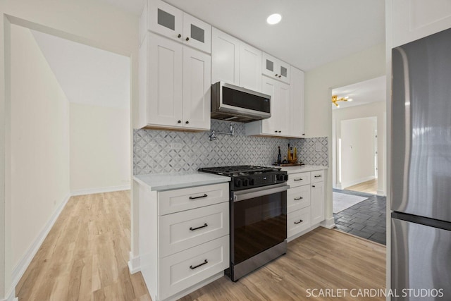 kitchen with tasteful backsplash, white cabinets, appliances with stainless steel finishes, glass insert cabinets, and light wood-type flooring