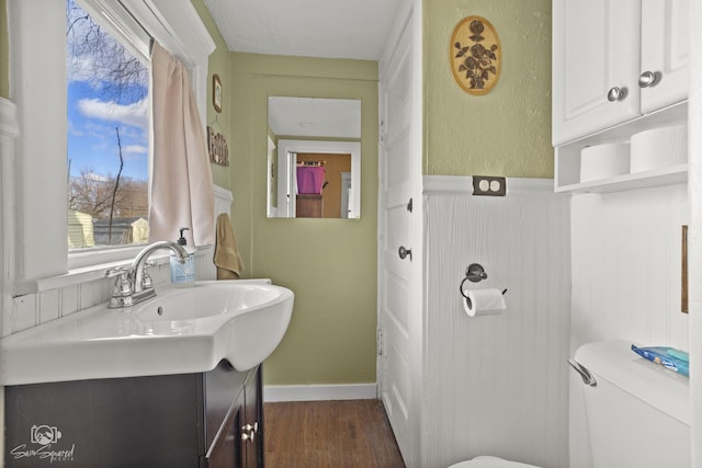 half bathroom with a textured wall, toilet, a wainscoted wall, wood finished floors, and vanity