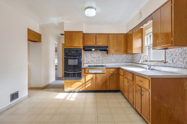 kitchen with brown cabinetry, light countertops, under cabinet range hood, a sink, and black oven