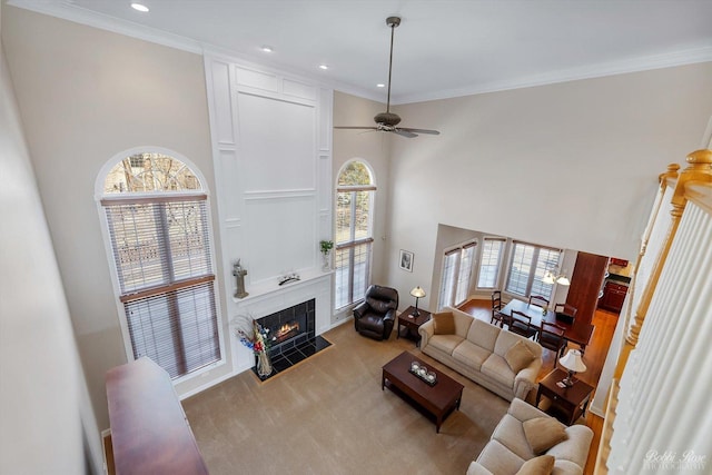 living area featuring a tiled fireplace, ornamental molding, carpet flooring, a high ceiling, and a ceiling fan