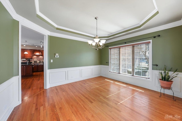 unfurnished dining area featuring a raised ceiling, a notable chandelier, wood finished floors, and wainscoting