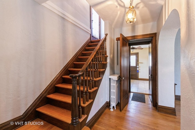 staircase featuring baseboards, arched walkways, a textured wall, radiator, and wood finished floors