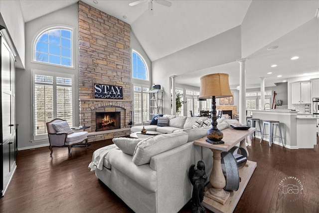 living room featuring ceiling fan, high vaulted ceiling, a stone fireplace, dark wood-style flooring, and decorative columns