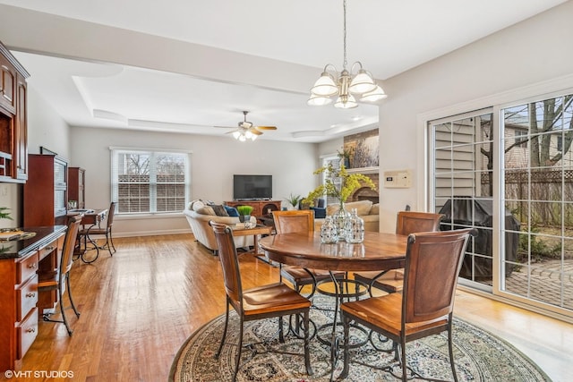 dining space featuring baseboards, a raised ceiling, light wood-style flooring, a fireplace, and ceiling fan with notable chandelier