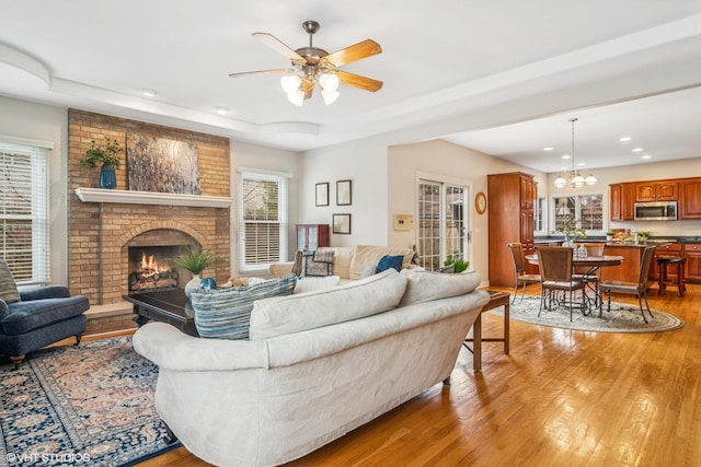 living area featuring recessed lighting, ceiling fan with notable chandelier, a fireplace, light wood-type flooring, and a raised ceiling