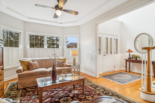 living room featuring light wood-type flooring, a raised ceiling, a ceiling fan, and baseboards