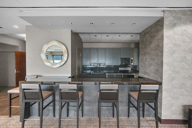 kitchen with gray cabinetry, a sink, appliances with stainless steel finishes, dark countertops, and a kitchen bar