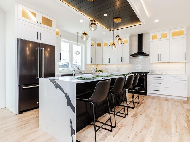 kitchen featuring a tray ceiling, high end fridge, wall chimney range hood, an island with sink, and black range