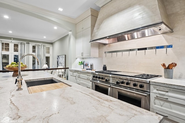 kitchen with range with two ovens, ornamental molding, a sink, light stone countertops, and wall chimney exhaust hood