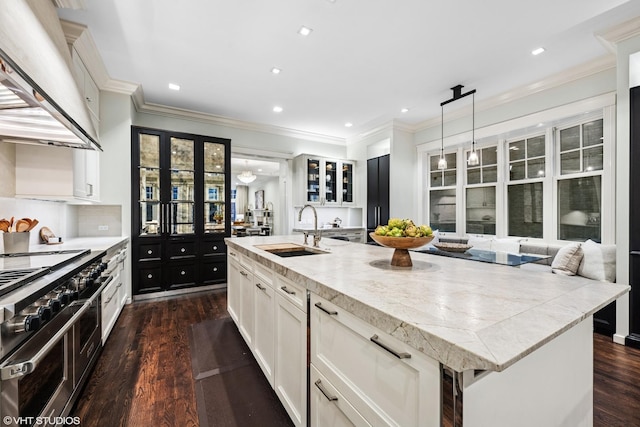 kitchen with a sink, range hood, double oven range, an island with sink, and dark wood finished floors