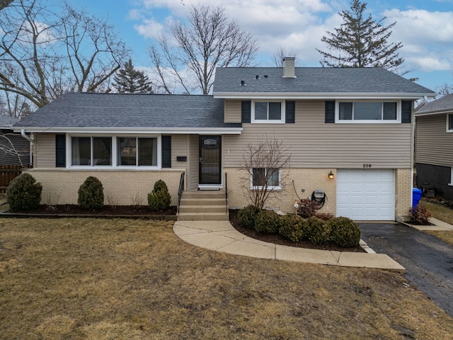 split level home featuring aphalt driveway, an attached garage, brick siding, roof with shingles, and a chimney