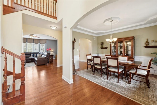 dining area with arched walkways, ceiling fan with notable chandelier, wood finished floors, ornamental molding, and a raised ceiling