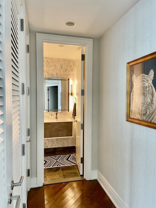 hallway with dark wood-style flooring, a sink, baseboards, and wallpapered walls