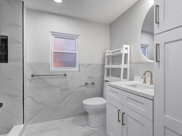 full bathroom featuring tiled shower, toilet, marble finish floor, vanity, and tile walls