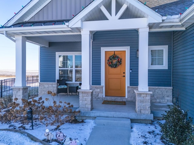 snow covered property entrance featuring stone siding, a shingled roof, a porch, and board and batten siding