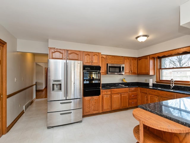 kitchen with dark stone countertops, brown cabinetry, a sink, appliances with stainless steel finishes, and a warming drawer