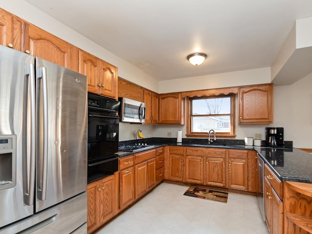 kitchen featuring brown cabinetry, dark stone counters, a sink, stainless steel appliances, and a warming drawer