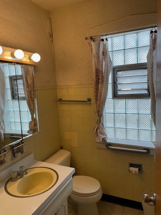 bathroom with toilet, a wainscoted wall, tile patterned flooring, vanity, and tile walls