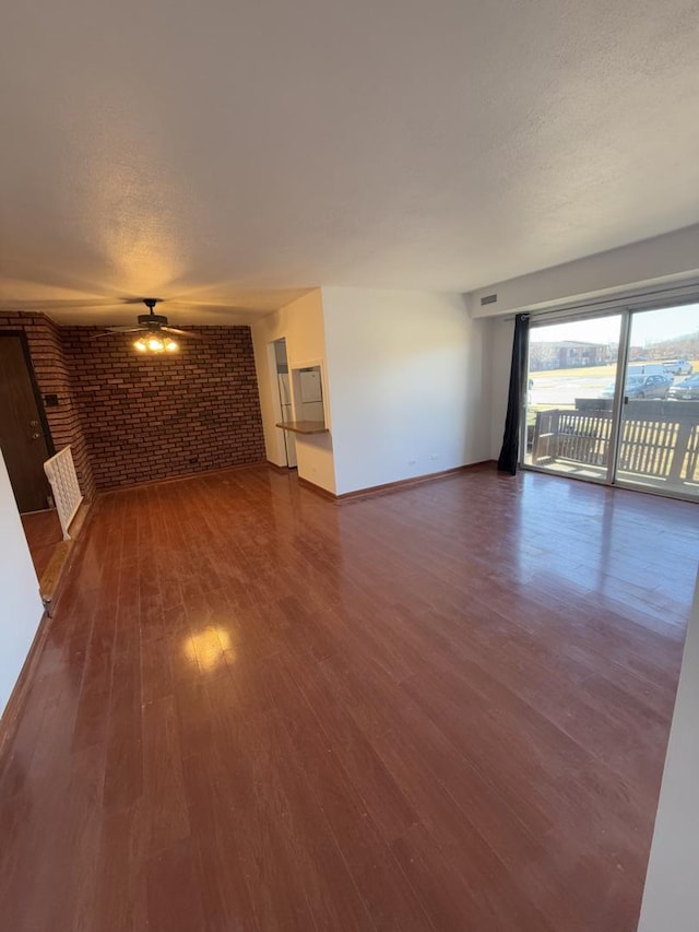 unfurnished living room with dark wood-style flooring, a ceiling fan, a textured ceiling, brick wall, and baseboards