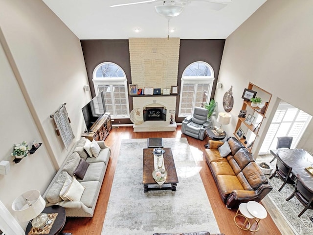 living area featuring a ceiling fan, a brick fireplace, a towering ceiling, and wood finished floors