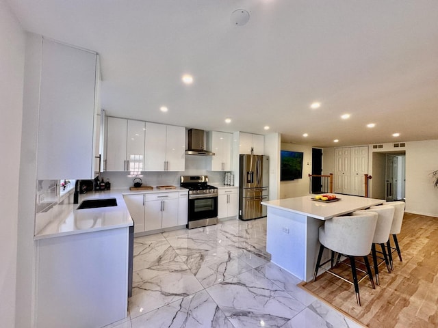 kitchen with white cabinets, stainless steel appliances, wall chimney range hood, a kitchen bar, and a sink