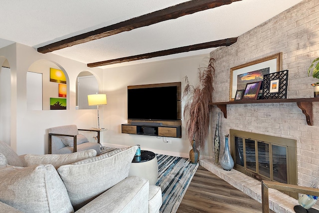 living room featuring a textured ceiling, wood finished floors, baseboards, a brick fireplace, and beamed ceiling